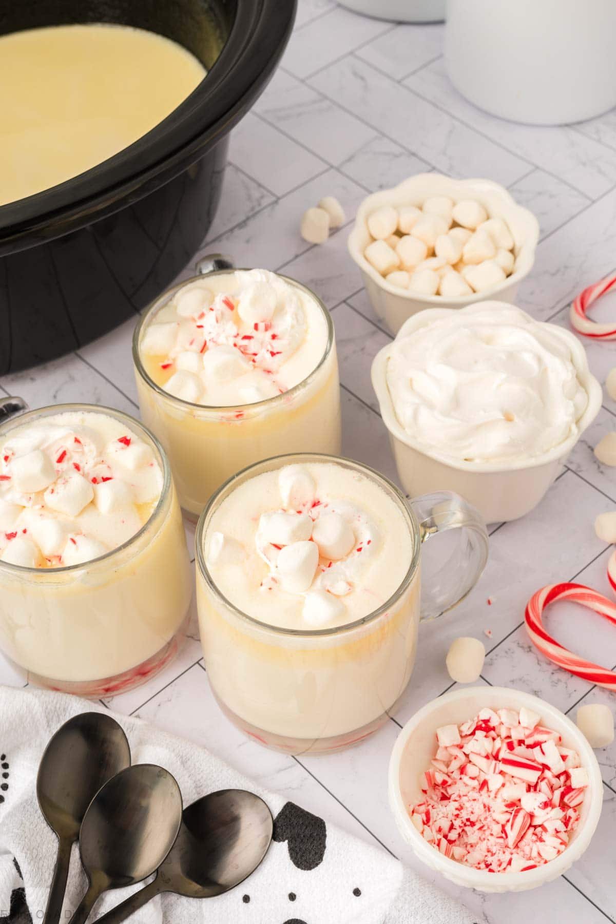 Three clear mugs of crock pot white hot chocolate recipe, topped with marshmallows and crushed peppermint, sit on a white surface surrounded by whipped cream, mini marshmallows, candy canes, spoons, and a black slow cooker.
