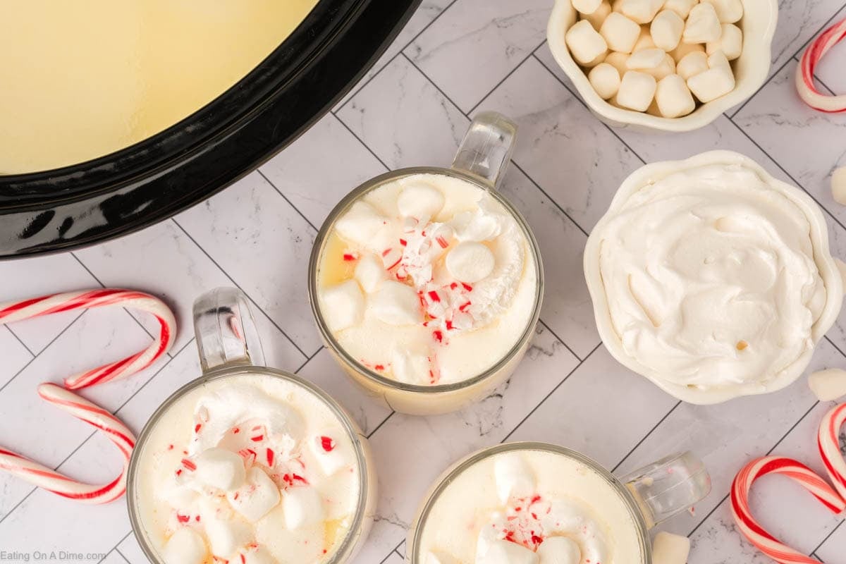 Three mugs of crock pot white hot chocolate recipe topped with whipped cream, mini marshmallows, and crushed candy canes sit on a white tiled surface, surrounded by candy canes, a bowl of marshmallows, and a bowl of whipped cream.
