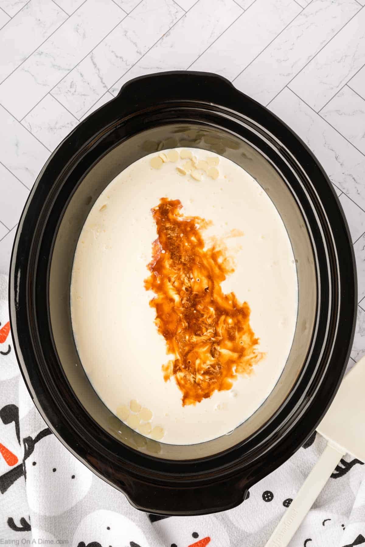 A crock pot white hot chocolate recipe simmers in the slow cooker, filled with cream, shredded cheese, and a splash of hot sauce, sitting on a white towel with black and orange designs atop a tiled surface.