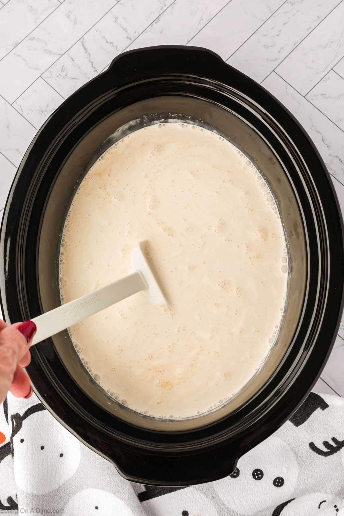 A hand is stirring a creamy white liquid in a black crock pot with a white spatula, making a delicious white hot chocolate recipe. The slow cooker sits on a white tiled surface next to a towel with a ghost pattern.