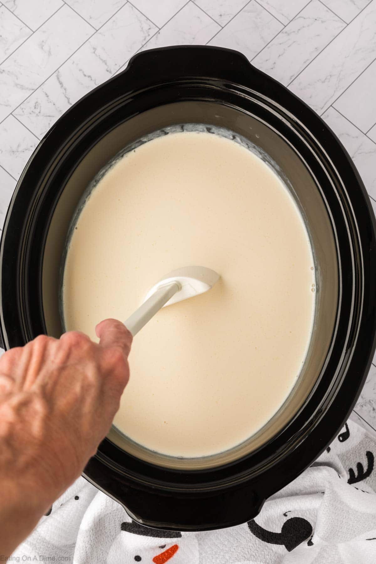 A hand stirs a creamy white liquid in a black crock pot, making a delicious white hot chocolate recipe. The cooker sits on a towel with a white tile floor in the background.