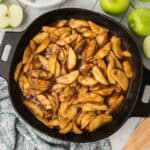 Sliced apples cooked in a dark, cinnamon-spiced sauce fill a black skillet, surrounded by fresh green apples and a patterned cloth on a light wooden surface—an inviting scene for this Easy Fried Apples Recipe.