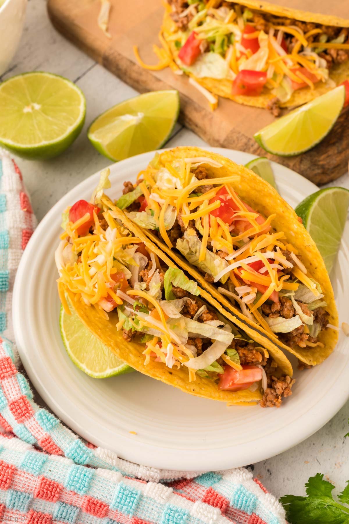 Ground Beef Tacos on a white plate topped with lettuce, shredded cheese and diced tomatoes. 