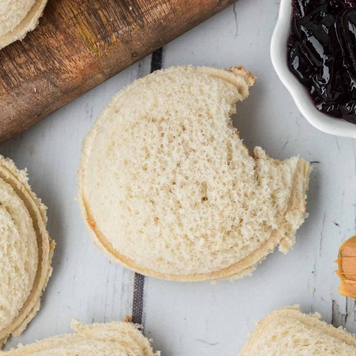 A round, crustless sandwich with a bite taken out of it, placed on a white wooden surface. To the right is a small dish of dark purple jam, and part of a wooden rolling pin is visible in the top-left corner. Other homemade uncrustables are partially visible at the edges.