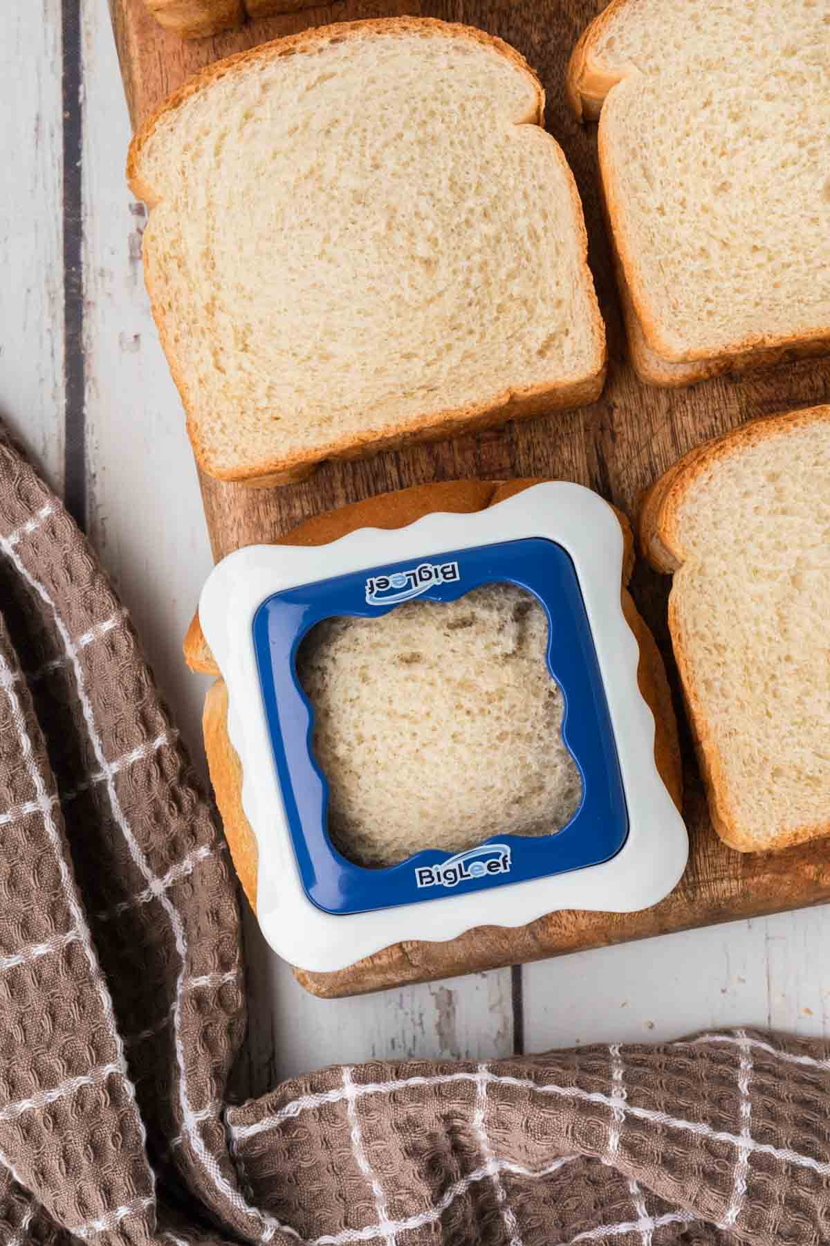 A sandwich cutter with a blue frame and white borders is placed on top of a slice of bread, surrounded by additional bread slices on a wooden surface. A brown and white checkered cloth is partially visible to the left side of the image, perfect for making homemade uncrustables effortlessly.