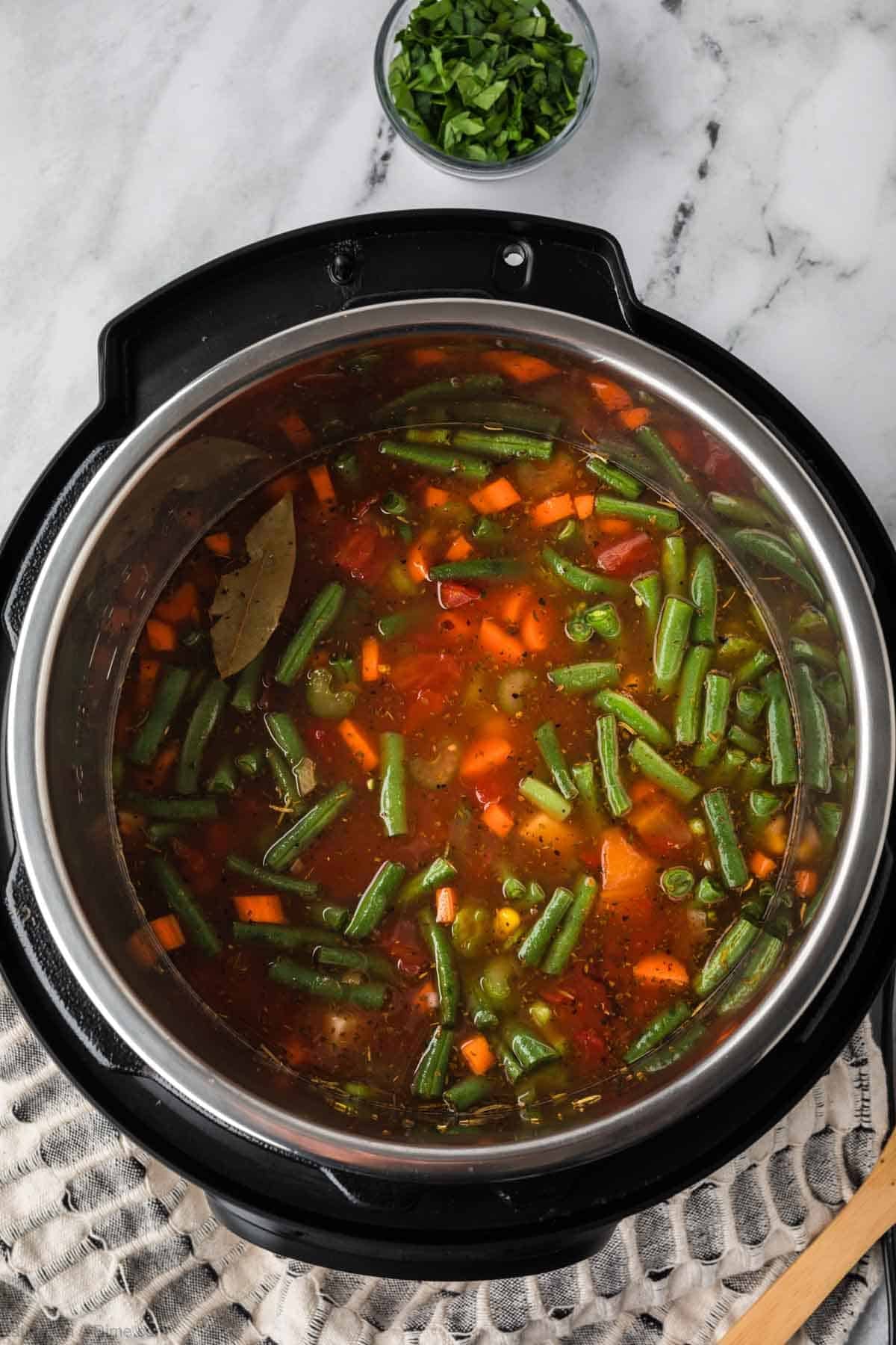 An overhead view of a pot filled with Instant Pot Vegetable Soup, featuring green beans, carrots, peas, and a bay leaf, set on a marble surface with chopped herbs in a small bowl nearby.