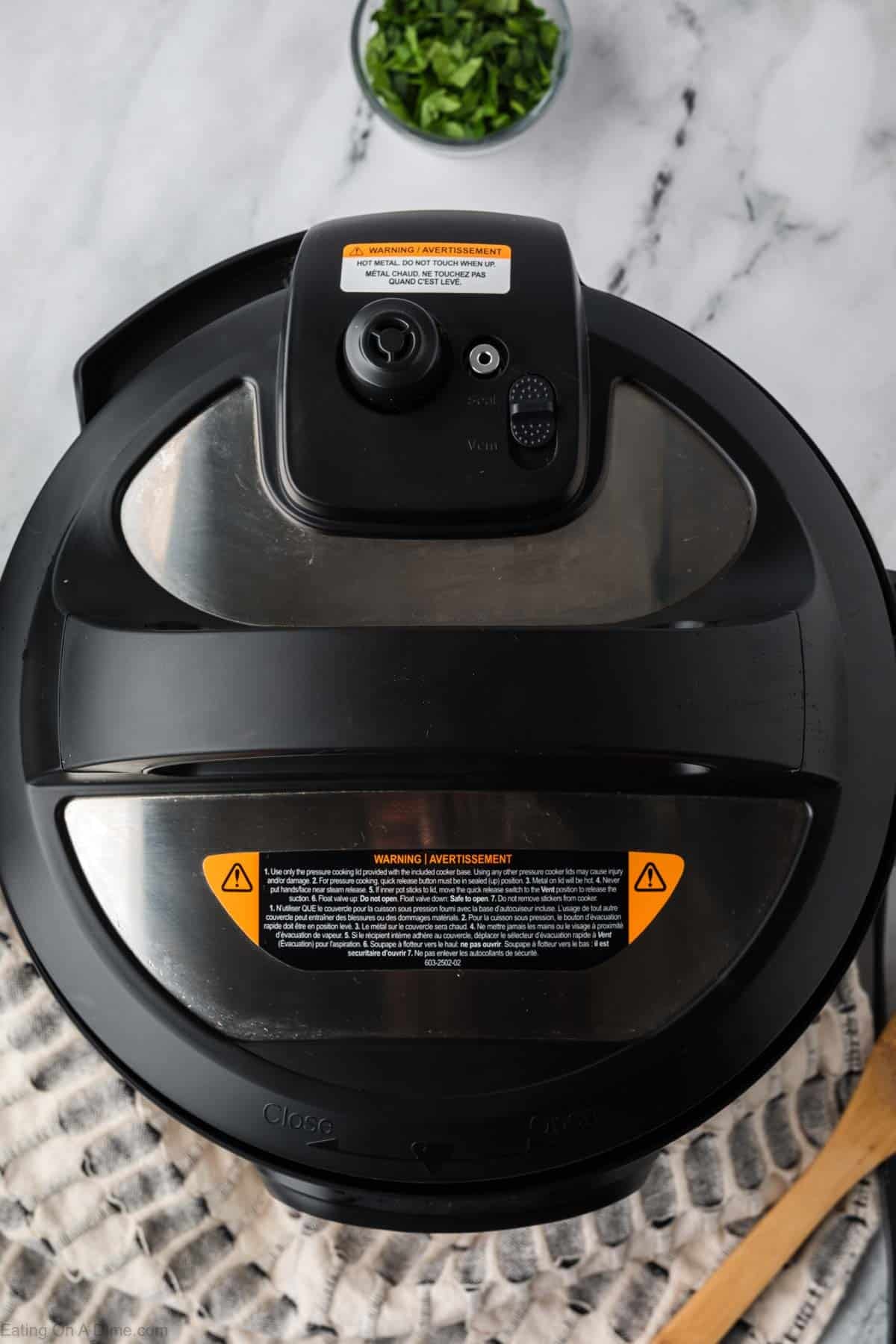 A close-up, top-down view of an Instant Pot lid on a kitchen counter hints at a fresh batch of Instant Pot Vegetable Soup, with a small bowl of chopped herbs and part of a wooden spoon visible nearby.