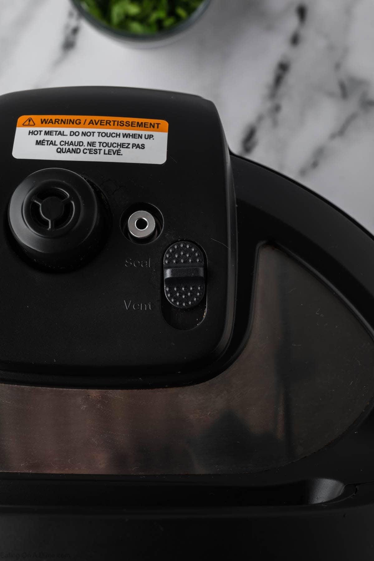 Close-up of the lid of a black pressure cooker, showing a warning label and a steam release valve with “Seal” and “Vent” settings—ready to make Instant Pot Vegetable Soup—on a marble countertop with a bowl of chopped herbs in the background.