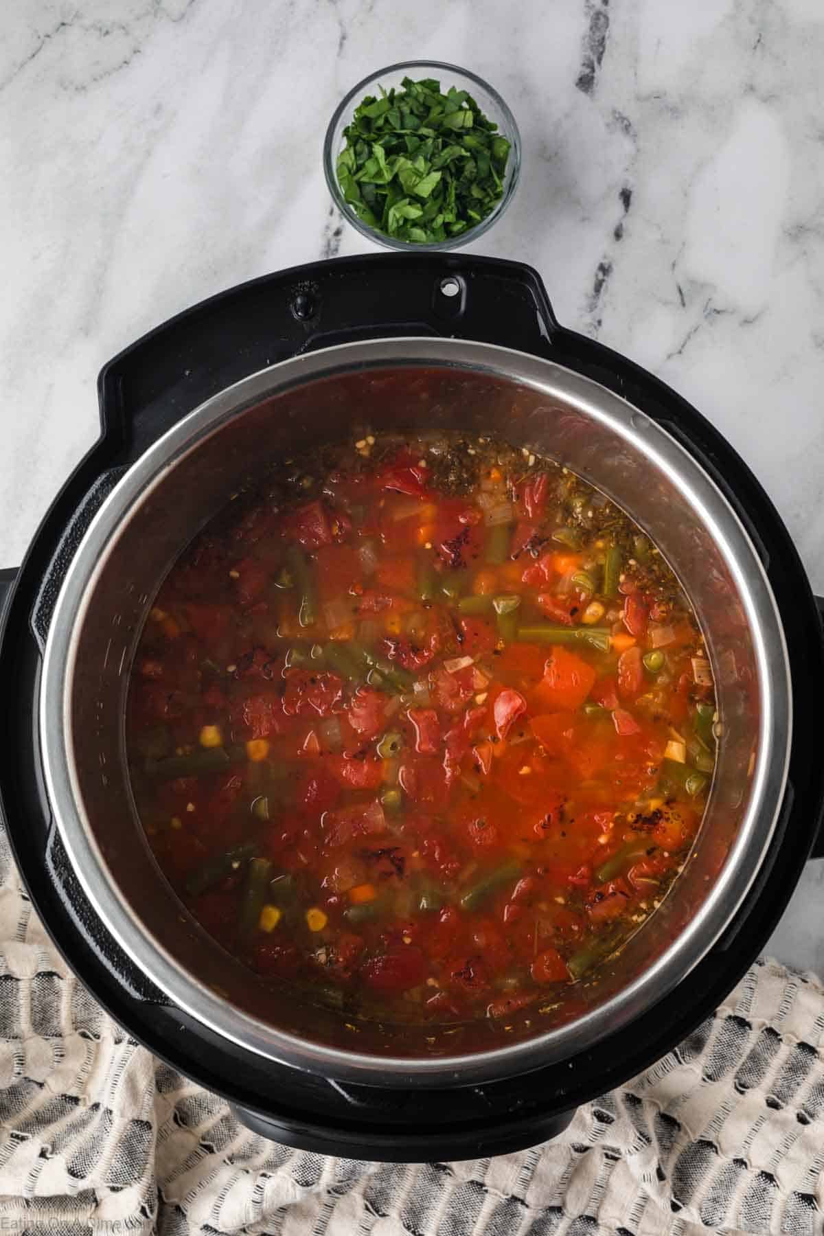 A pot of Instant Pot Vegetable Soup with tomatoes, green beans, corn, and herbs sits on a marble countertop. Above it is a small bowl of chopped fresh parsley, while a textured towel rests nearby.