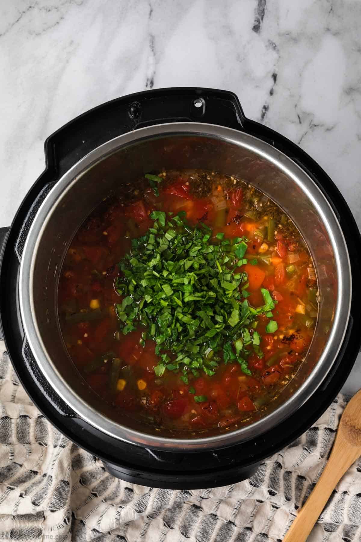 A pot of hearty Instant Pot Vegetable Soup, topped with freshly chopped green herbs, sits on a marble countertop beside a wooden spoon and a patterned cloth.