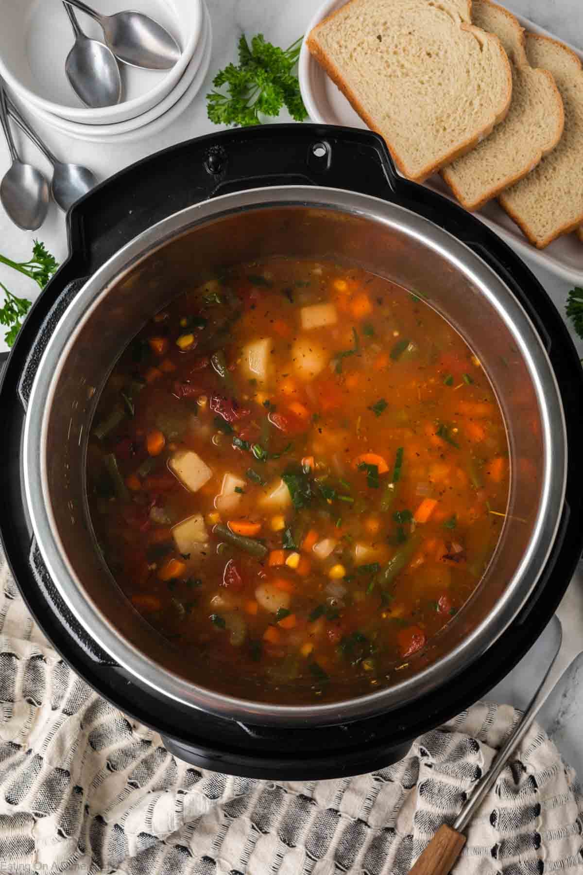 A pot of Instant Pot Vegetable Soup with carrots, potatoes, and herbs sits in an Instant Pot. Surrounding the soup are a plate of sliced bread, a stack of bowls, and spoons on a marble countertop.