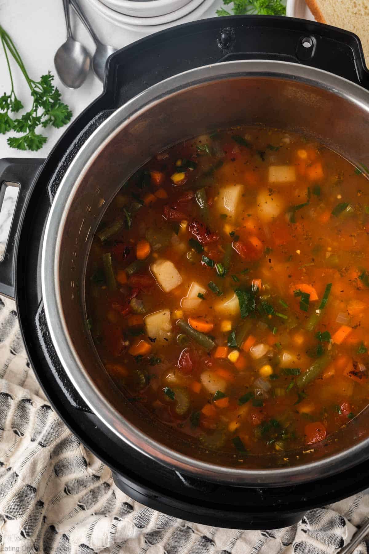 A pot of hearty Instant Pot Vegetable Soup with potatoes, carrots, corn, tomatoes, and green beans sits in an Instant Pot on a patterned kitchen towel, with fresh parsley and spoons nearby.