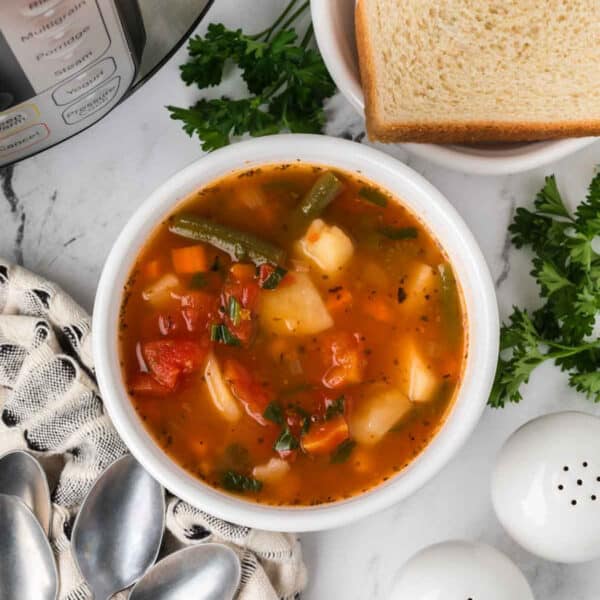 A bowl of Instant Pot Vegetable Soup with potatoes, green beans, and tomatoes sits on a marble counter next to slices of bread, fresh parsley, a patterned napkin, spoons, and salt and pepper shakers.