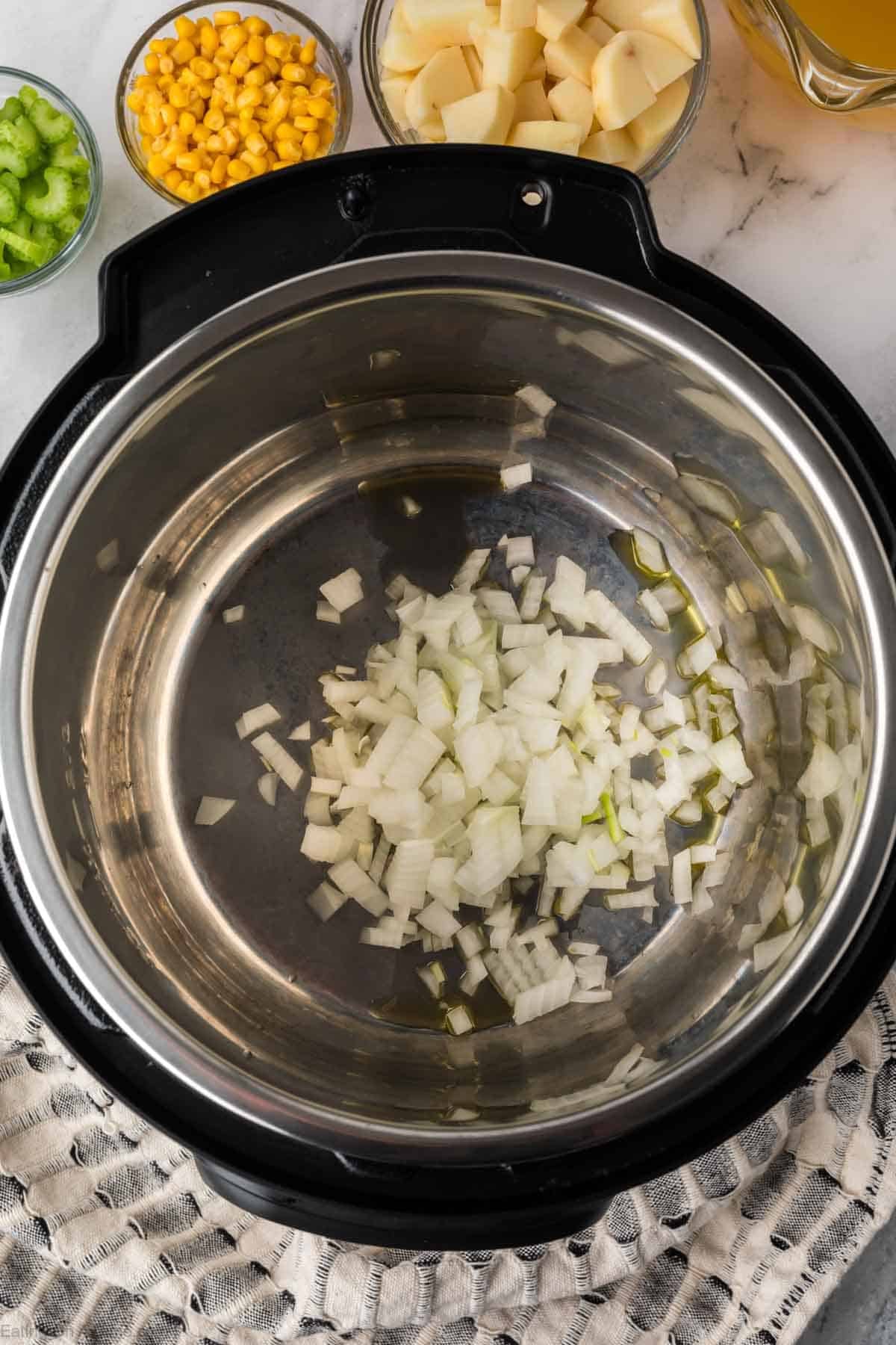 Chopped onions are cooking in an Instant Pot for a hearty Instant Pot Vegetable Soup. Nearby on the marble countertop are bowls of diced potatoes, corn, chopped celery, and a bowl with yellow broth ready to be added.