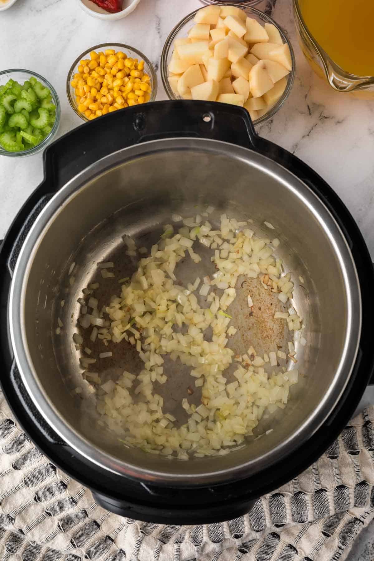 Chopped onions sautéing in an Instant Pot for a hearty Instant Pot Vegetable Soup. Surrounding the pot are bowls of diced potatoes, corn, celery, and a pitcher of broth, all arranged on a patterned cloth.