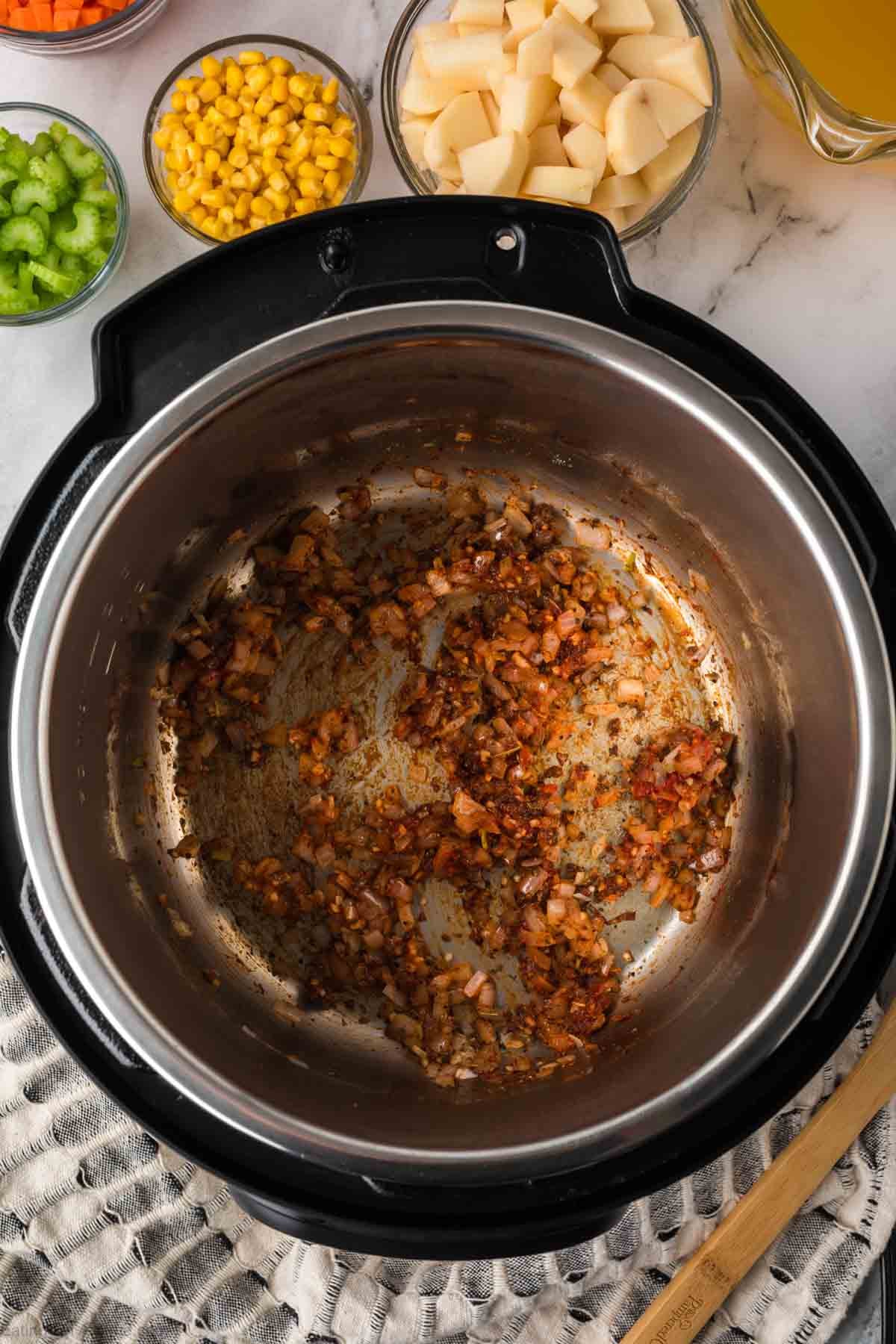 An Instant Pot Vegetable Soup in progress, with sautéed onions and seasonings at the bottom, surrounded by small bowls of diced potatoes, corn, celery, and a measuring cup of broth on a marble countertop.