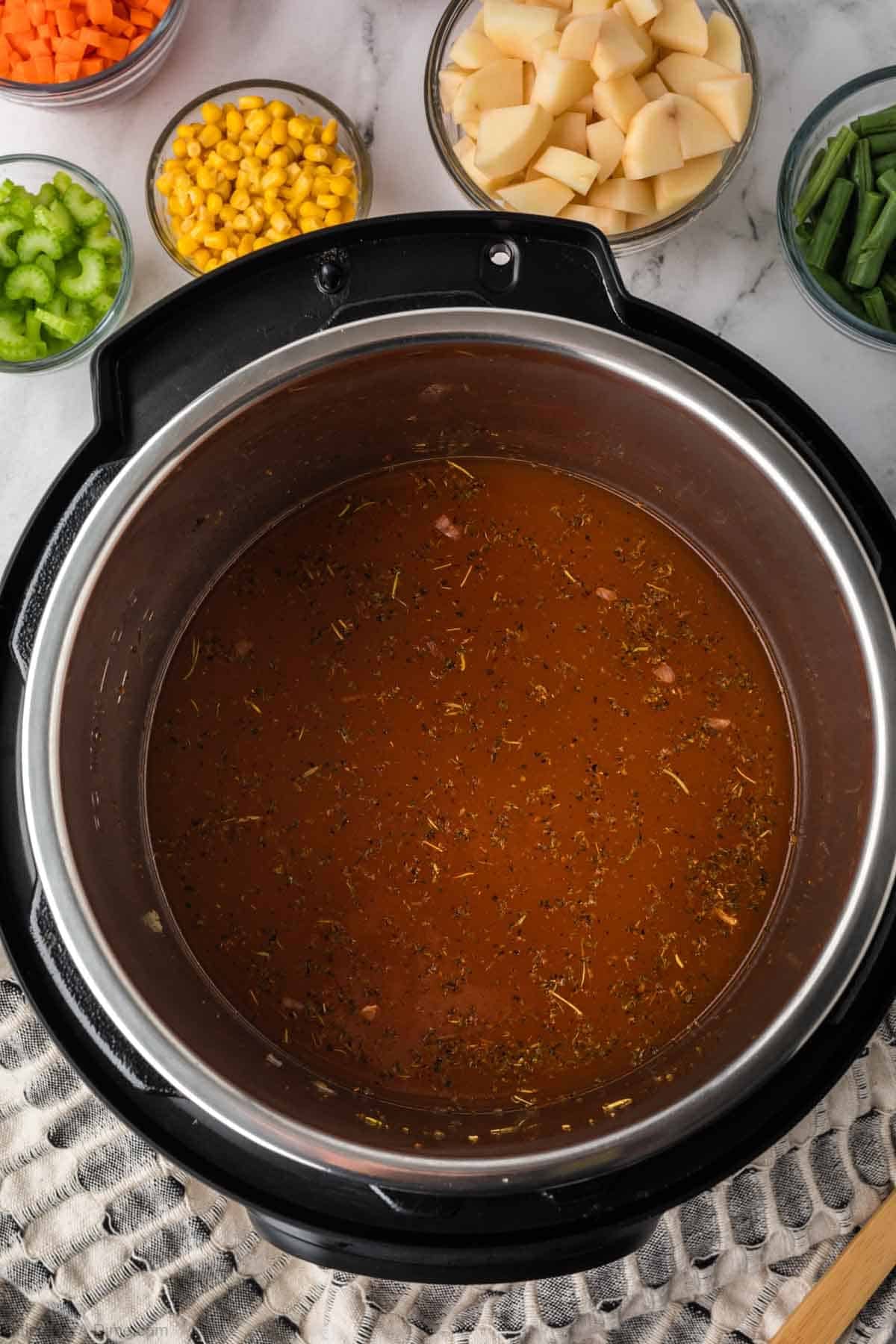 An Instant Pot filled with seasoned broth sits on a countertop, surrounded by bowls of chopped potatoes, carrots, celery, corn, and green beans—everything you need to make a hearty Instant Pot Vegetable Soup.