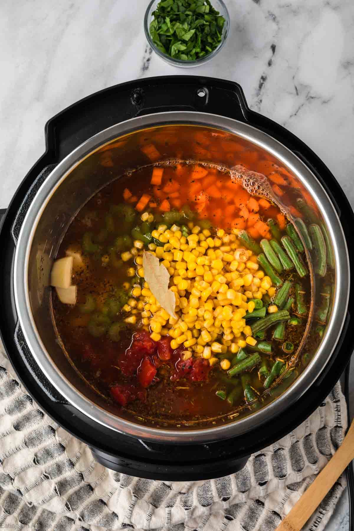 A top-down view of Instant Pot Vegetable Soup filled with broth, corn, carrots, green beans, tomatoes, celery, potatoes, and a bay leaf sits on a trivet, with a bowl of chopped herbs nearby.