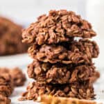 A close-up of a stack of four chocolate oatmeal cookies on a white surface, with more no bake pretzel cookies blurred in the background.