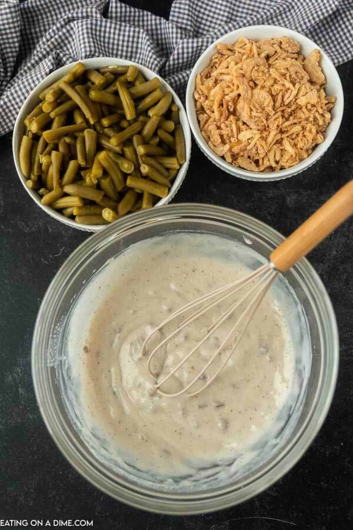 Combining the cream of mushroom soup, milk, and spices in a bowl with a whisk with small bowls of green beans and French fried onions nearby.