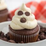 A Ghost Cupcake with white frosting shaped like a spooky spirit, decorated with chocolate chips for eyes and mouth, sits in a bowl of chocolate chips. A red and white cloth is blurred in the background.