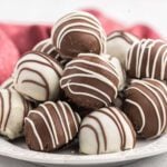 A plate of Oreo Truffles coated in white and milk chocolate, drizzled with contrasting chocolate, arranged in a pile with a red cloth in the background.