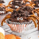 An Easy Spider Cupcake decorated with candy eyes, pretzel stick legs, and crumbled cookie pieces on top, sits in a shiny silver wrapper on a cooling rack.