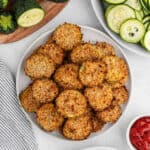A white plate filled with crispy Air Fryer Zucchini Chips, alongside fresh sliced zucchini and whole zucchini, set on a light-colored surface.