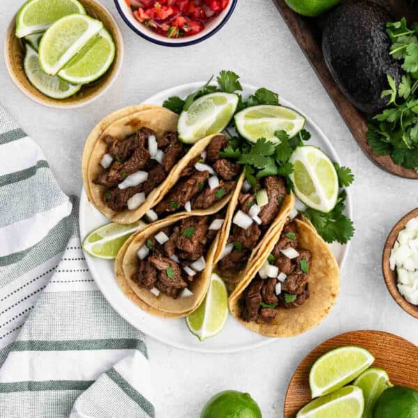 3 Carne Asada Tacos on a white plate topped with chopped onions with a fresh cilantro and lime wedges.