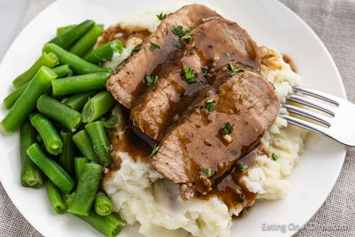 Sliced pork tenderloin over mashed potatoes with a side of green beans on a plate.