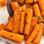 A close-up of this easy roasted carrots recipe shows carrot sticks on a plate, garnished with chopped parsley and black pepper, with a serving spoon visible.