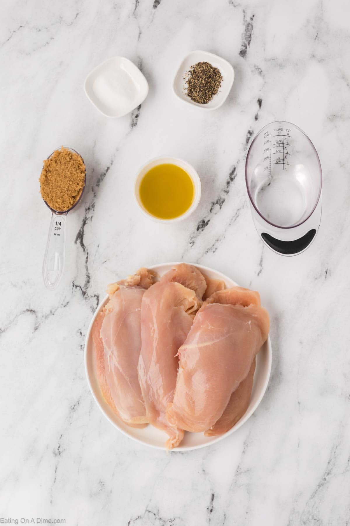 Raw chicken breasts on a white plate are ready to become Brown Sugar Chicken, surrounded by dishes of brown sugar, olive oil, salt, pepper, and a measuring cup with liquid on a white marble surface.