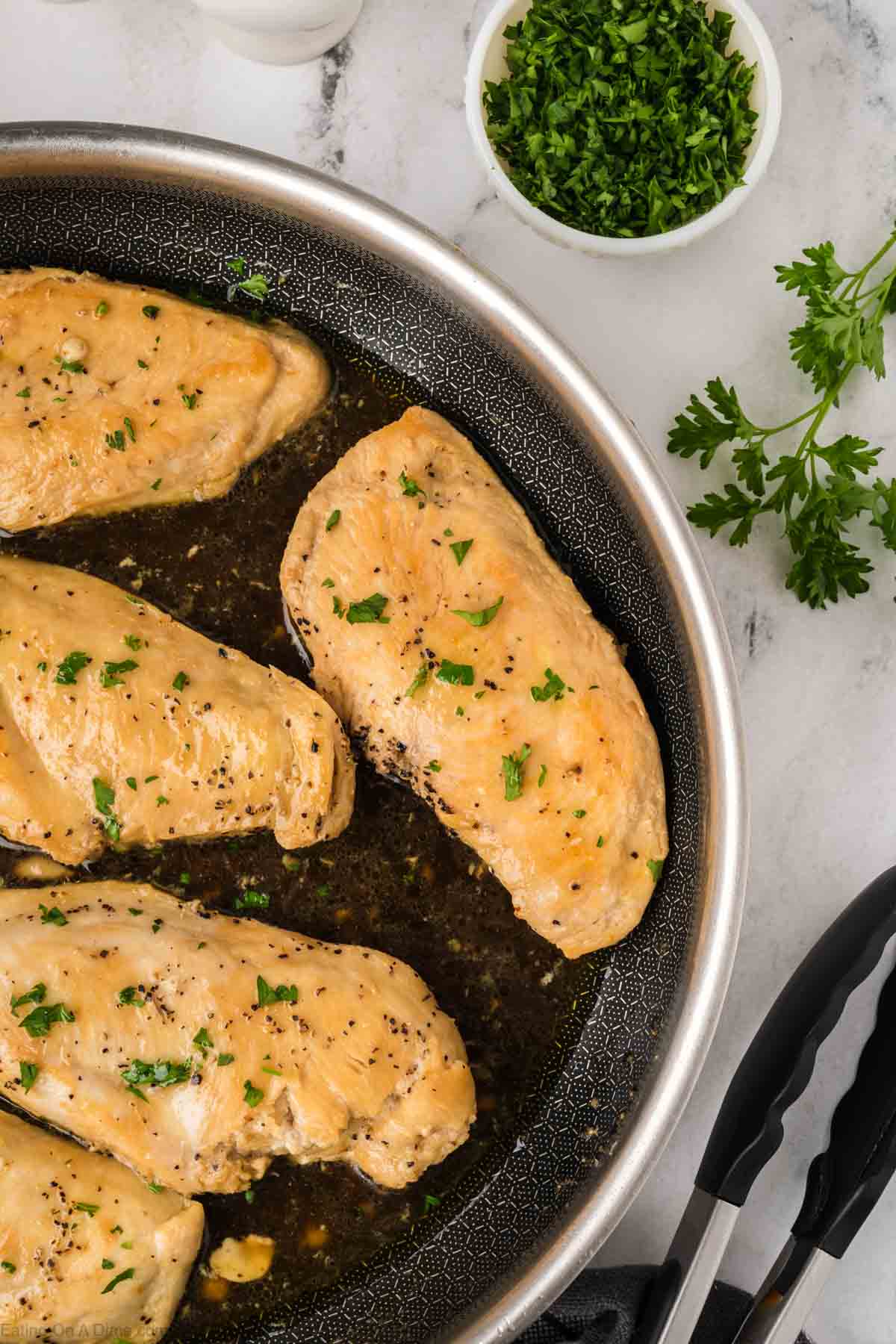 A skillet filled with Brown Sugar Chicken breasts garnished with chopped parsley, next to a bowl of fresh parsley and a pair of tongs on a marble countertop.