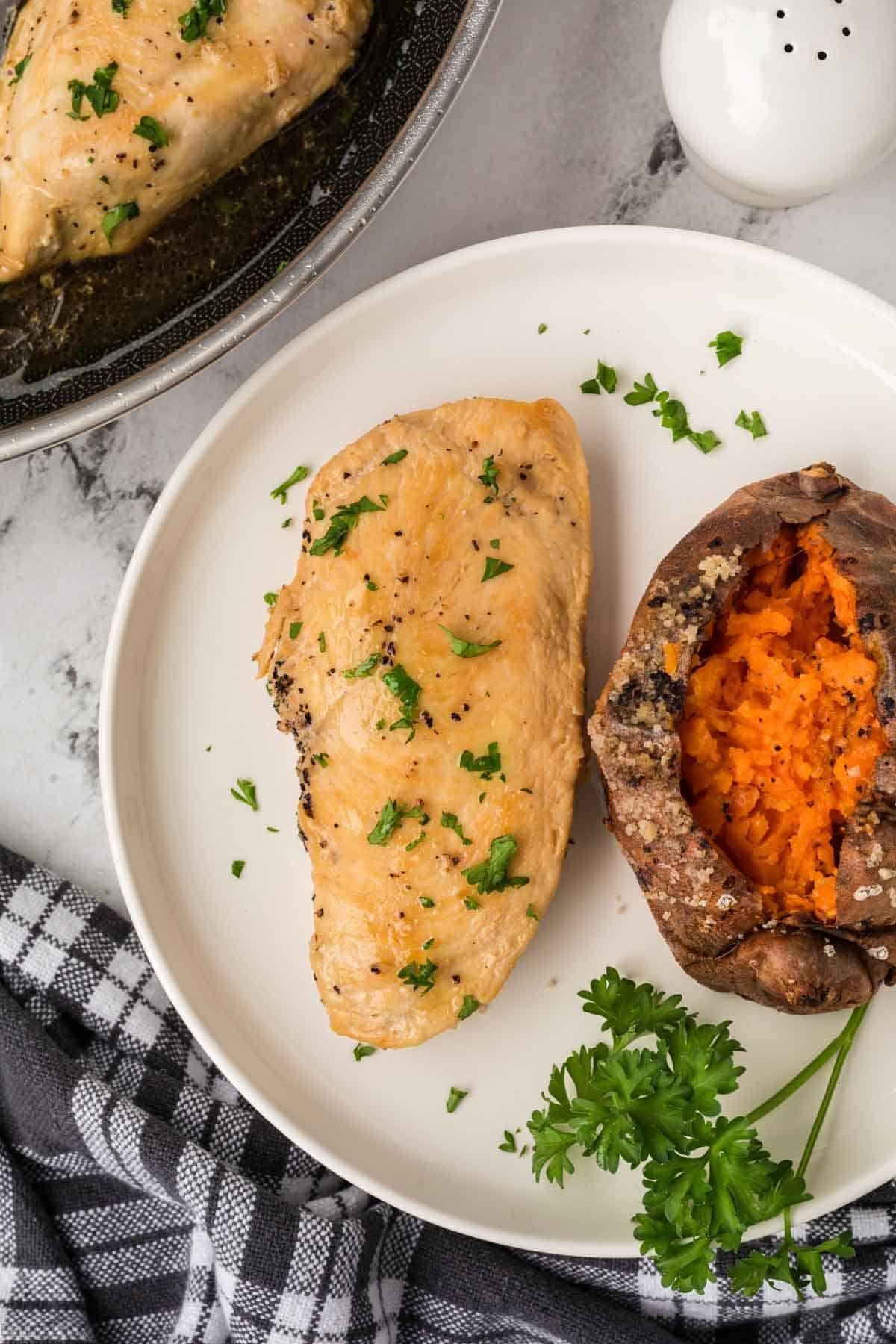 A plate with a Chicken breast garnished with parsley and a baked sweet potato, next to a sprig of fresh parsley. A pan with another chicken breast and a black and white towel are visible nearby.
