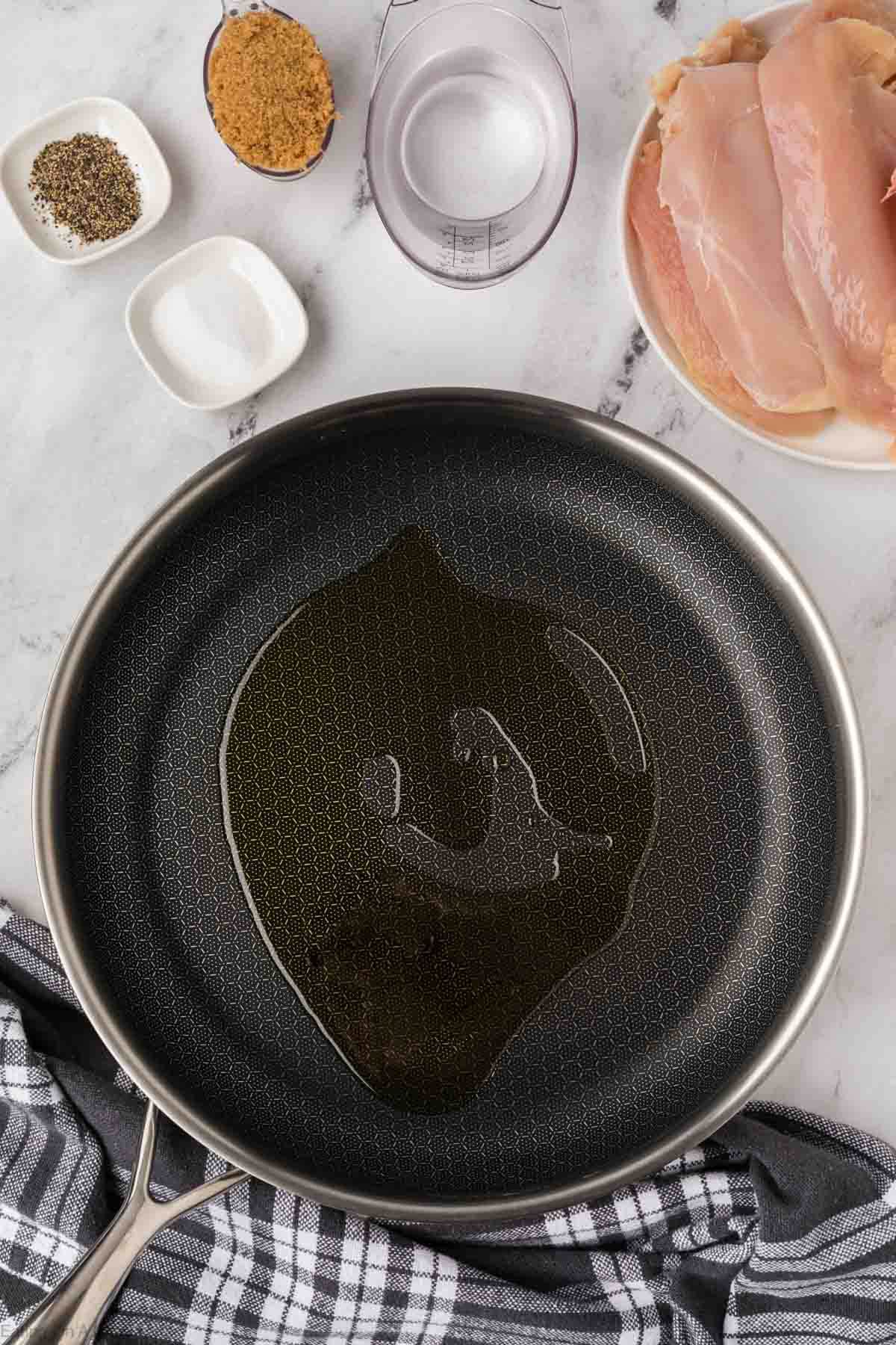 A frying pan with oil sits on a marble surface, surrounded by ingredients for Brown Sugar Chicken: a plate of raw chicken breasts, a cup of water, bowls of brown sugar, salt, pepper, and a gray-striped kitchen towel.