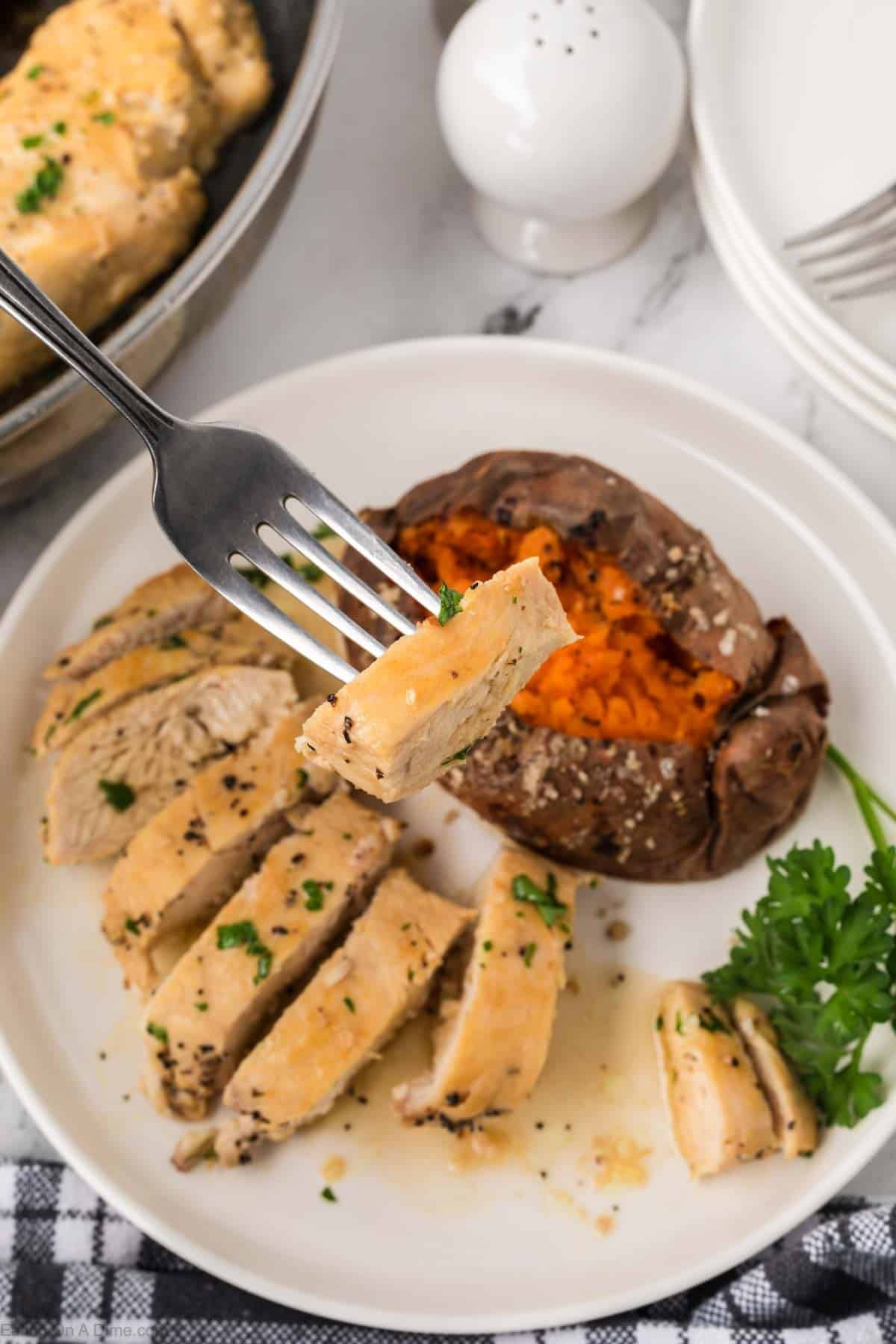 A plate with sliced Brown Sugar Chicken breast garnished with herbs, a baked sweet potato sprinkled with salt, and a sprig of parsley. A fork holds a piece of chicken above the plate.