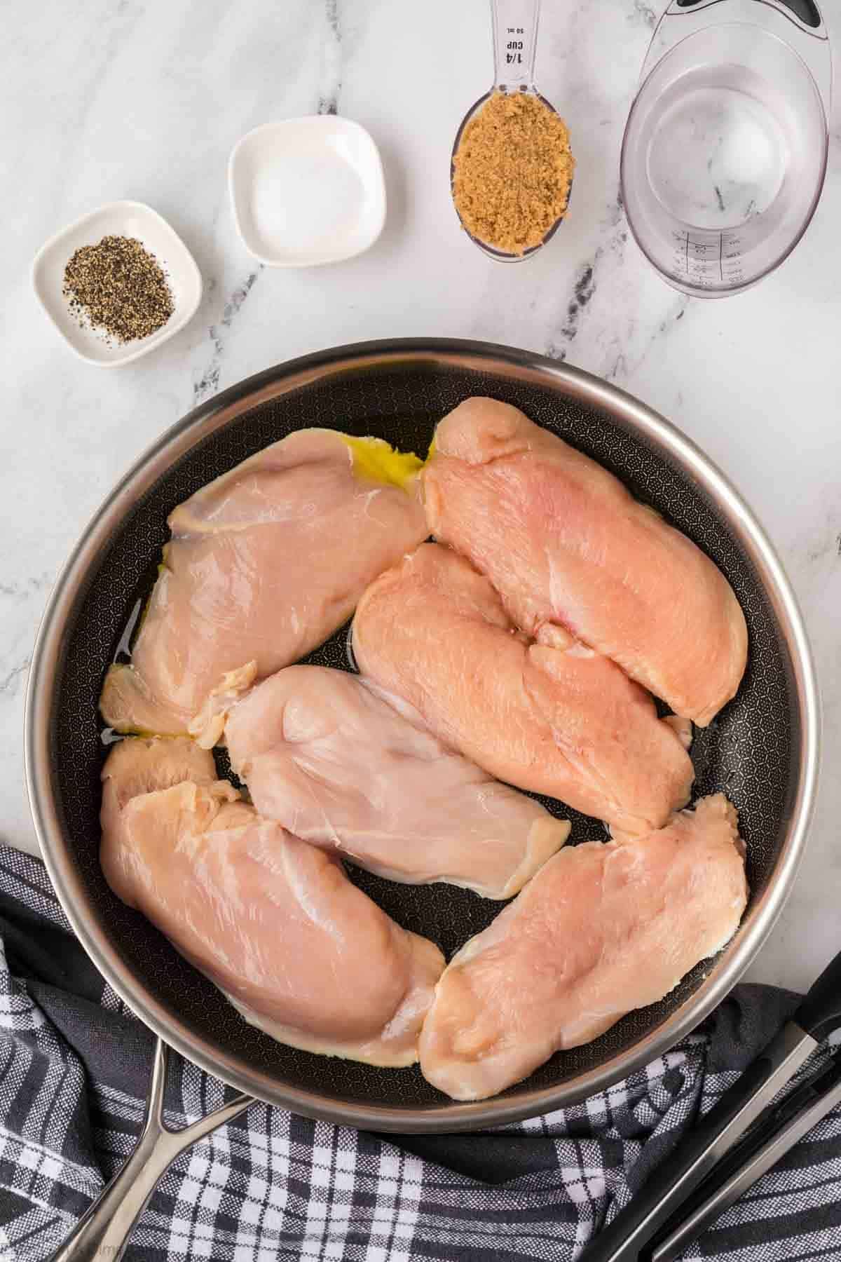 Six raw chicken breasts are arranged in a large frying pan on a marble countertop, with bowls of black pepper, salt, and brown sugar—perfect for making Brown Sugar Chicken—a measuring cup of water, and a striped towel nearby.
