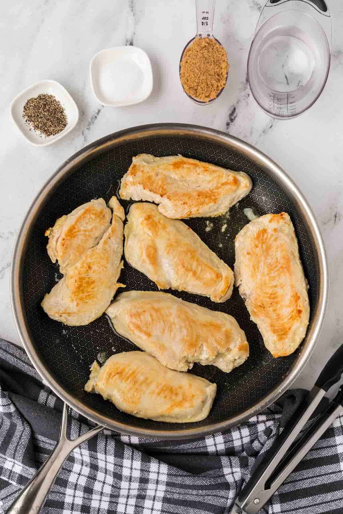 A skillet with six browned Brown Sugar Chicken breast pieces on a stovetop, surrounded by small bowls of black pepper, salt, brown sugar, a measuring cup of water, and a black-and-white striped kitchen towel.