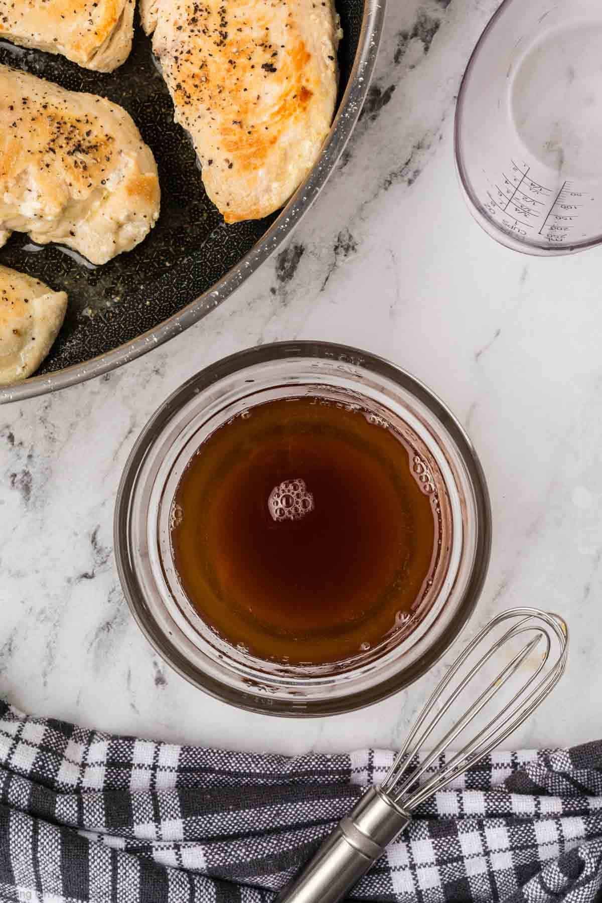 A bowl of brown liquid for Brown Sugar Chicken sits on a marble countertop with a whisk beside it. Nearby, cooked seasoned chicken breasts are in a pan, along with a measuring cup and a black-and-white towel.
