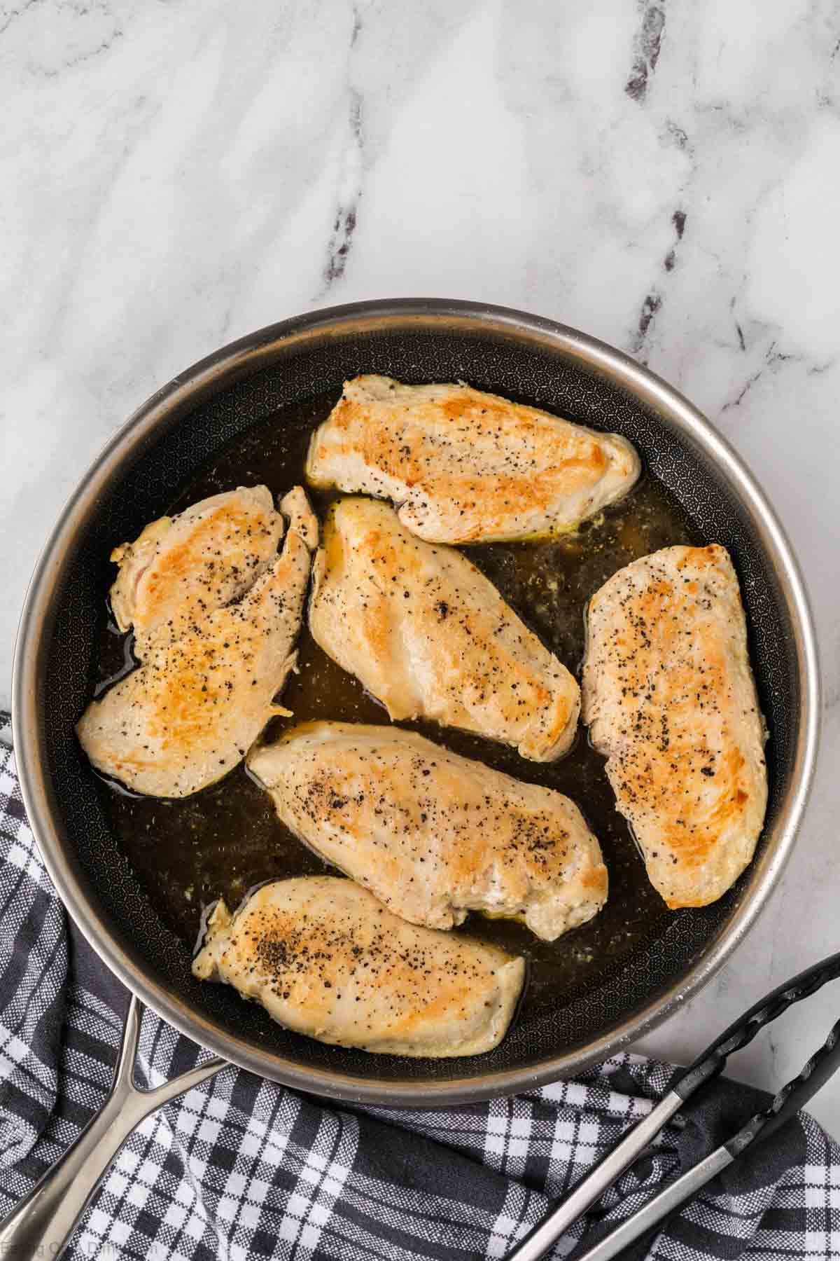 Six cooked Brown Sugar Chicken breast pieces seasoned with pepper are arranged in a stainless steel skillet. The skillet rests on a white marble surface next to a black-and-white checkered towel and black kitchen tongs.