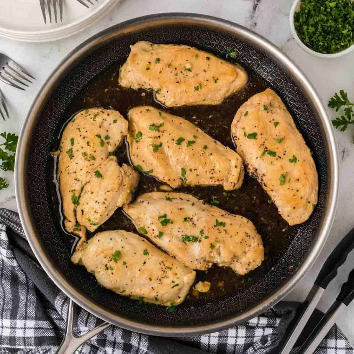 Six cooked Brown Sugar Chicken breasts garnished with chopped parsley are arranged in a frying pan with a dark sauce. The pan sits on a marble surface next to a black-and-white napkin, forks, stacked plates, and fresh parsley.