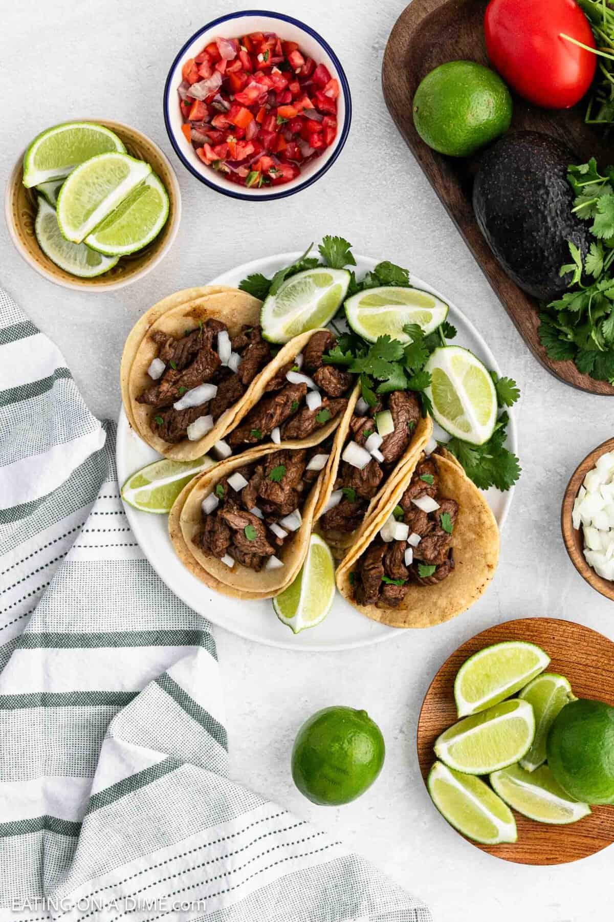 4 small street tacos topped with chopped onions and fresh cilantro on a plate with a side of fresh lime.