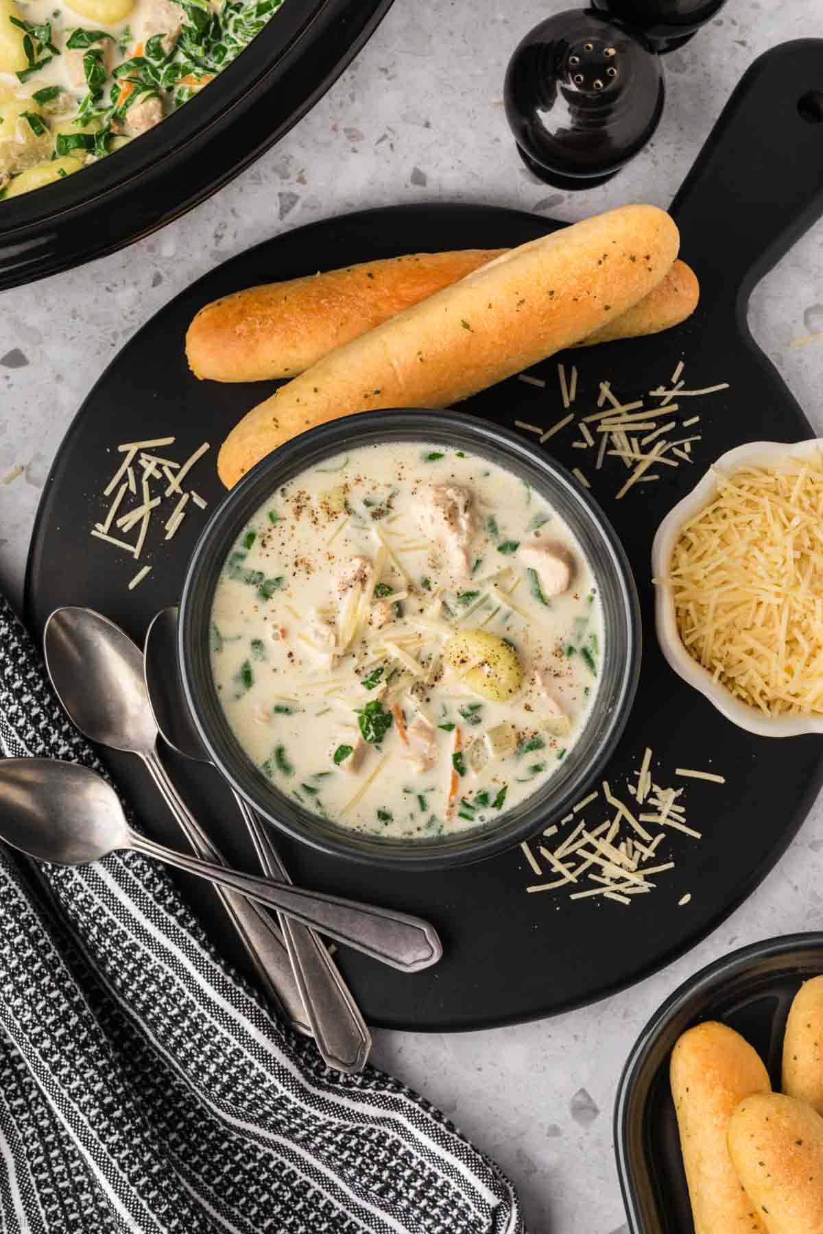 Chicken Gnocchi Soup in a black bowl on a platter with shredded parmesan cheese and breadsticks