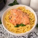 A bowl of cooked spaghetti topped with slow cooker tomato basil chicken, covered in a creamy pink herb sauce and garnished with fresh basil. Two forks, a patterned napkin, and two jars sit nearby on a white surface.