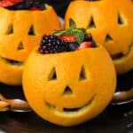 Three Jack-O-Lantern Oranges carved with Halloween faces are filled with fresh berries and displayed on a dark plate, creating a festive and healthy Halloween snack.