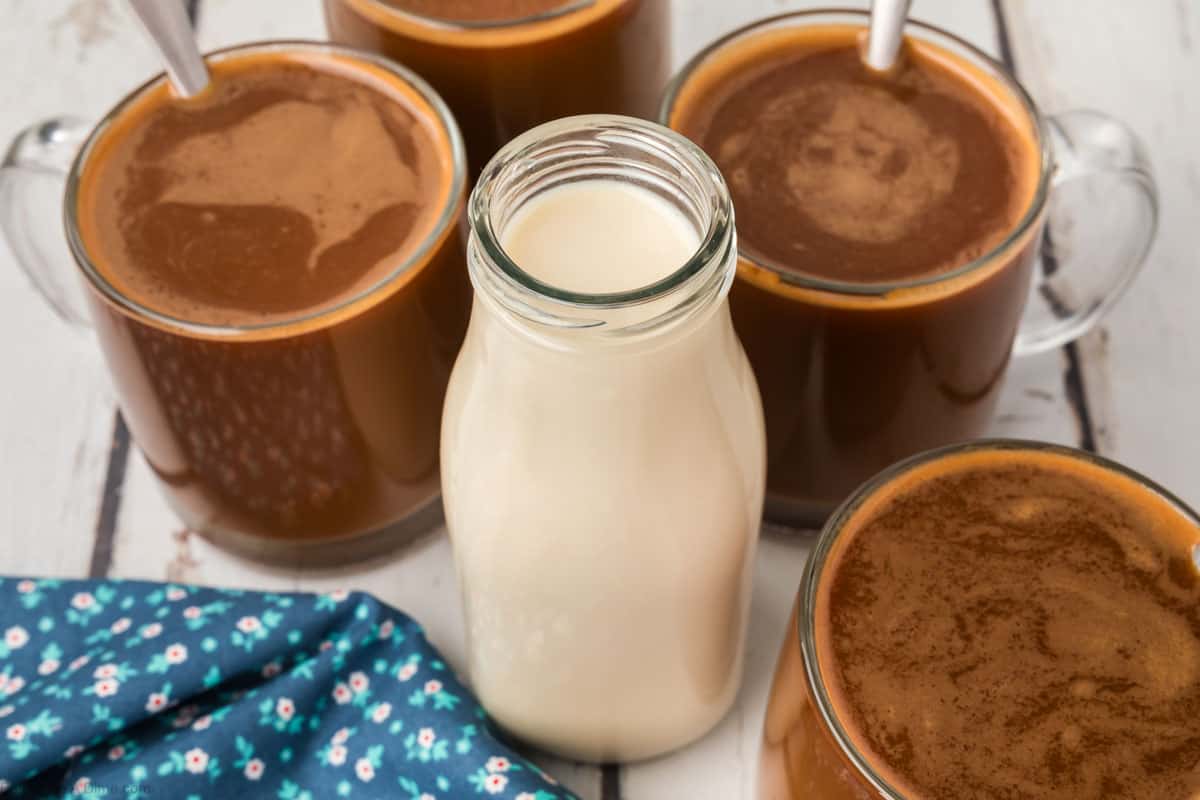 A glass milk bottle is centered on a table, surrounded by five cups filled with frothy coffee. There is a blue napkin with a floral pattern placed to the left. The combination of milk and coffee hints at the addition of homemade coffee creamer for an extra touch of flavor.