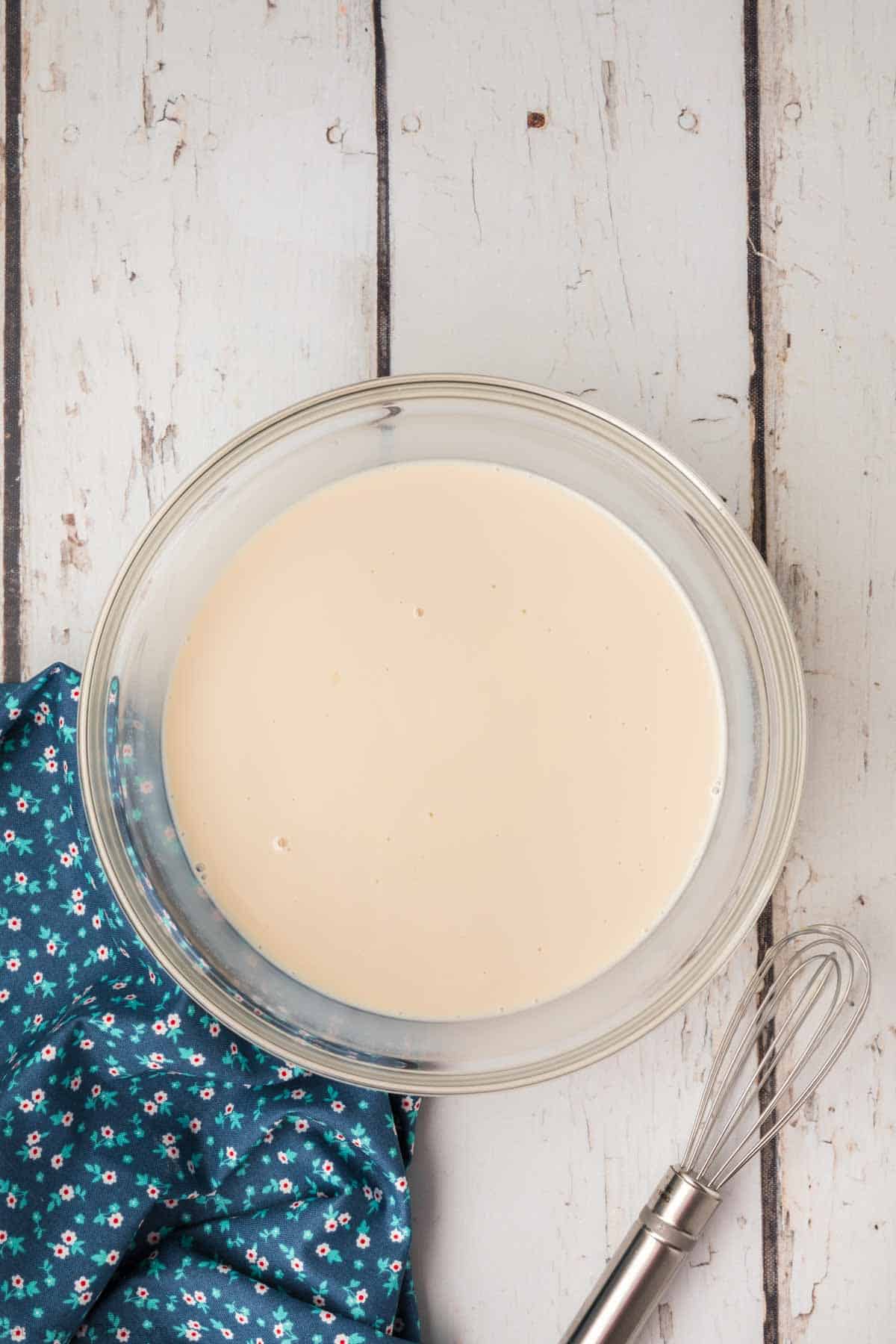 A glass bowl filled with smooth, creamy batter sits on a white wooden surface. A silver whisk with a metallic handle rests nearby. A folded blue cloth with small white and pink floral patterns lies adjacent to the bowl and whisk, ready for making homemade coffee creamer.
