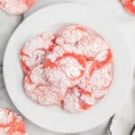 A white plate filled with round, pink Strawberry cake mix Cookies coated in powdered sugar, arranged on a marble surface. The cookies have a crackled texture and are grouped together in the center of the plate.