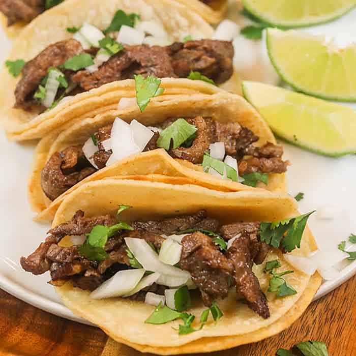 3 Steak street tacos topped with white onions and cilantro on a white plate with lime wedges on the right. 
