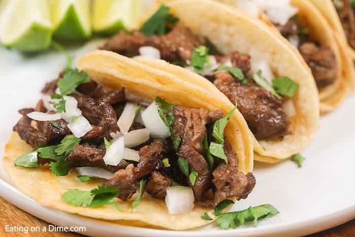 4 beef street tacos on a white plate topped with white onions and cilantro.  Each taco is on 2 corn tortillas. 